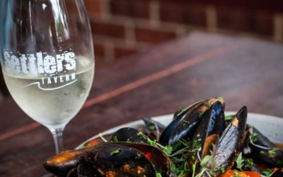 Settlers_Tavern_Food_OBJ887376 A bowl of mussels in tomato sauce with herbs next to a glass of white wine on a wooden table.