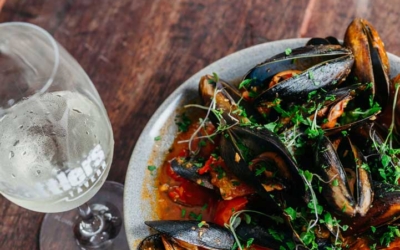 Settlers_Tavern_Food_OBJ887381 A plate of mussels in sauce with herbs, next to a glass of white wine on a wooden table.