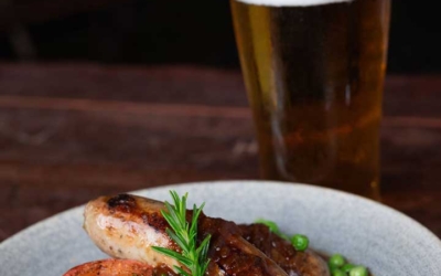 Settlers_Tavern_Food_OBJ887392 A plate of sausages with mashed potatoes, peas, and a grilled tomato slice, garnished with rosemary, next to a glass of beer.