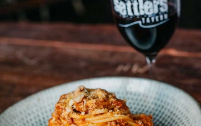 Settlers_Tavern_Food_OBJ887396 A plate of spaghetti bolognese topped with cheese is on a table next to a glass of red wine.
