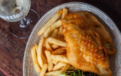 Settlers_Tavern_Food_OBJ887430 A plate of fried fish and chips with a side salad and lemon wedge, served with a dipping sauce. A glass of white wine is on the wooden table beside it.