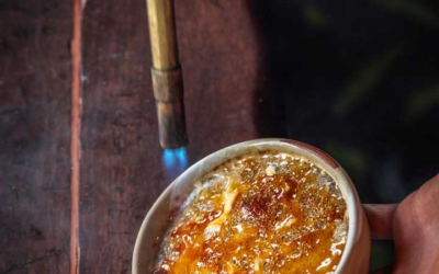 Settlers_Tavern_Food_OBJ887445 A hand uses a handheld torch to caramelize the sugar on top of a creme brulee in a bowl on a wooden surface.