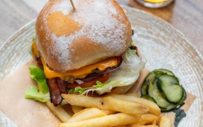 Settlers_Tavern_Food_OBJ887459 A cheeseburger with lettuce, tomato, and bacon on a bun, served with fries and pickle slices on a plate. A glass of beer is in the background.