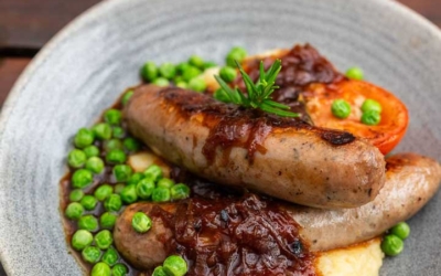 Settlers_Tavern_Food_OBJ887462 A plate of sausages with mashed potatoes, peas, grilled tomato, and onion gravy, garnished with rosemary on a wooden table.