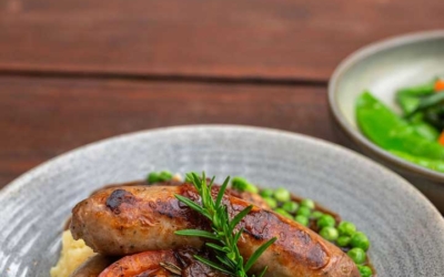 Settlers_Tavern_Food_OBJ887464 Plate with sausages, mashed potatoes, peas, and a slice of tomato, garnished with rosemary, on a wooden table.