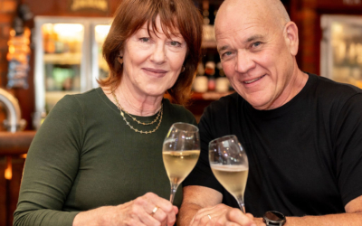 Settlers_Tavern_Food_OBJ887546 A man and woman smile while holding glasses of champagne at a bar, with bottles and decorations in the background.