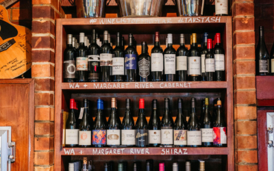 Settlers_Tavern_Food_OBJ887570 Wine bottles arranged on wooden shelves with handwritten labels indicating varieties like Margaret River Cabernet and Shiraz. Glassware displayed below, set against a brick wall in a cozy setting.