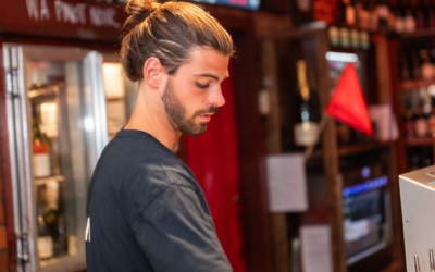 Settlers_Tavern_Food_OBJ887596 Man with a bun hairstyle behind a bar counter, preparing a drink. Bottles and a digital display are visible in the background.