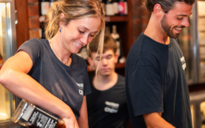Settlers_Tavern_Food_OBJ887604 Two bartenders in black shirts prepare drinks at a bar. Bottles of liquor and wine are visible on shelves behind them.