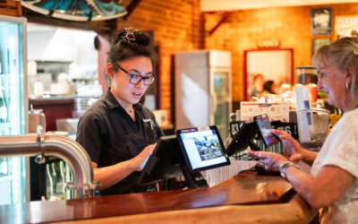 Settlers_Tavern_Food_OBJ887609 A restaurant employee in a black uniform uses a touchscreen at the counter, assisting a customer who is interacting with another screen. Kitchen equipment is visible in the background.