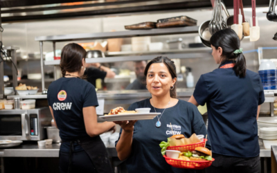 Settlers_Tavern_Food_OBJ887682 A restaurant employee holds plates of food in a busy kitchen with two other employees working in the background.
