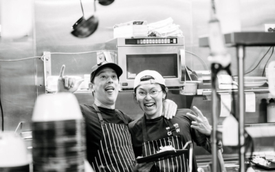 Settlers_Tavern_Food_OBJ887693 Two people wearing aprons and caps, smiling and posing in a busy kitchen with stacked plates and hanging ladles.