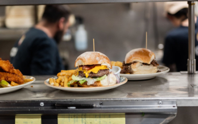 Settlers_Tavern_Food_OBJ887696 Plates with burgers, fries, and condiments are ready on a restaurant kitchen pass. Chefs work in the blurred background.
