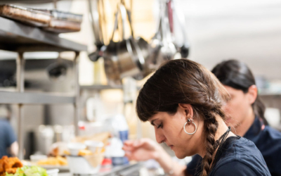 Settlers_Tavern_Food_OBJ887704 Two kitchen staff members are preparing food in a busy restaurant kitchen. One focuses on plating, while the other checks order tickets.
