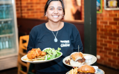 Settlers_Tavern_Food_OBJ887712 A person smiling while holding plates with a burger, fries, and salad in a restaurant setting.
