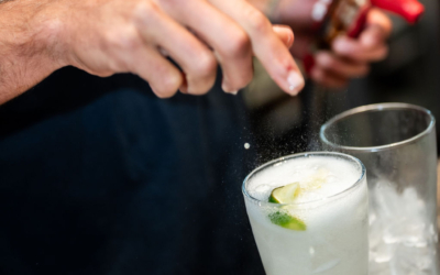 Settlers_Tavern_Food_OBJ887717 Person sprinkling salt into a glass of iced beverage garnished with lime slices, with a drink bottle in the background and a cloth on the counter.