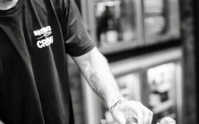 Settlers_Tavern_Food_OBJ887721 Bartender preparing a drink, pouring liquid from a jigger into a glass, in a bar setting. Black and white image.